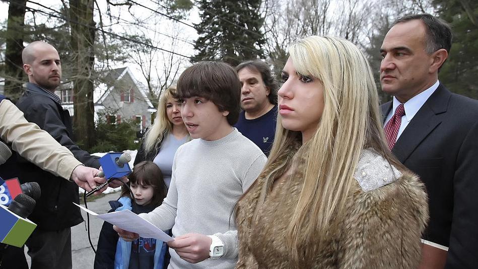 A group of people stands outdoors while a teenage boy reads from a paper as reporters hold microphones toward him.