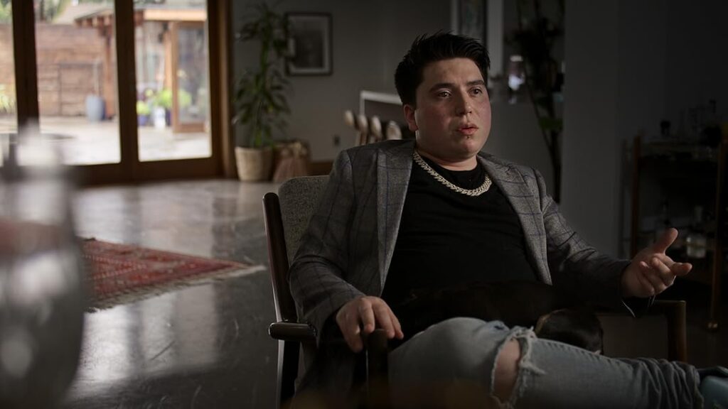 A man wearing a gray blazer, black shirt, chain necklace, and ripped jeans sits in a modern, dimly lit room with large windows and indoor plants, captured with ZEISS lens clarity.