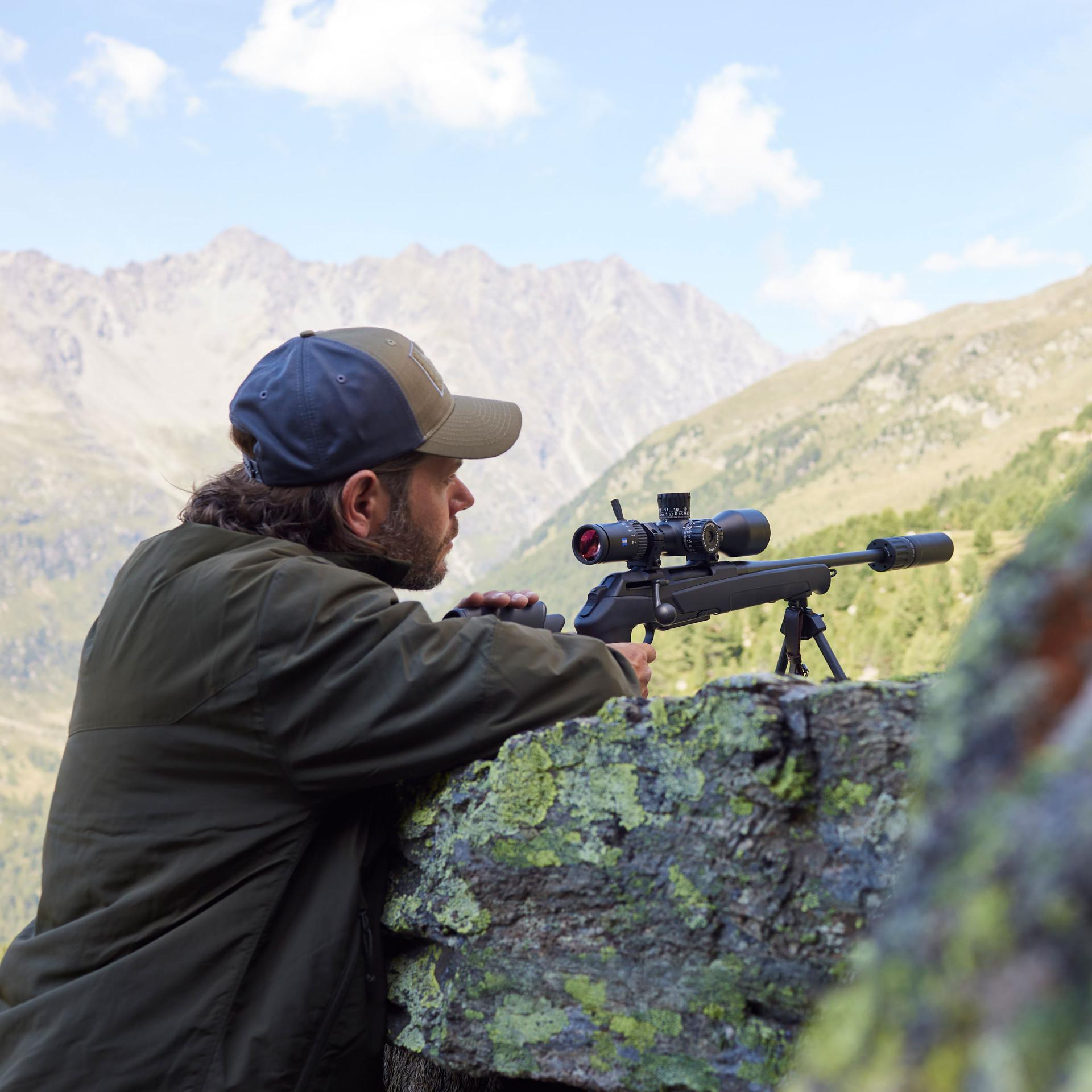 Hunter with a rifle mounted with the ZEISS LRP S3 636-56 riflescope, lying on rocks in the mountains – optimized for precise shots at very long distances