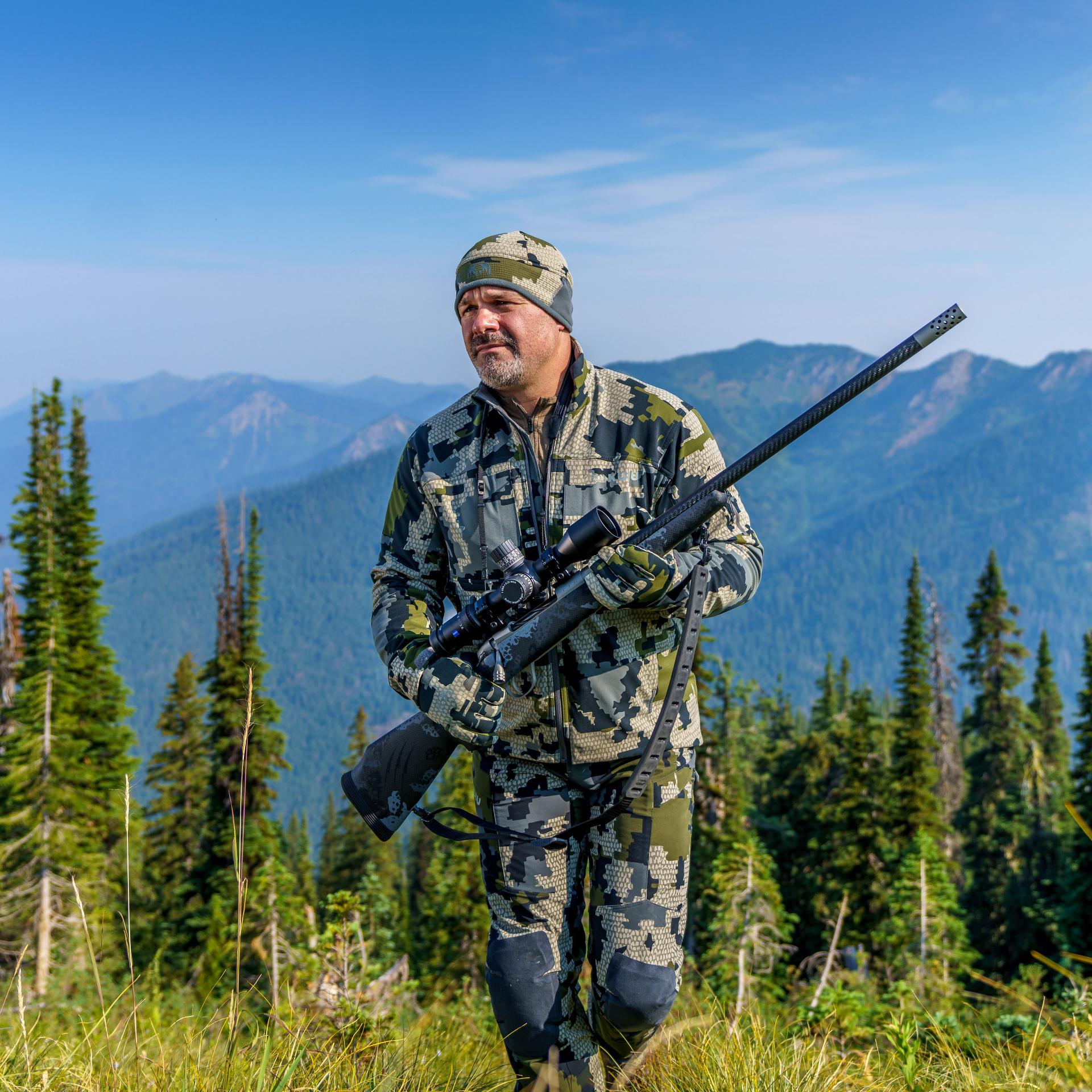 Hunter carrying a rifle equipped with a ZEISS LRP S5 525-56 riflescope while walking through a forested mountain landscape
