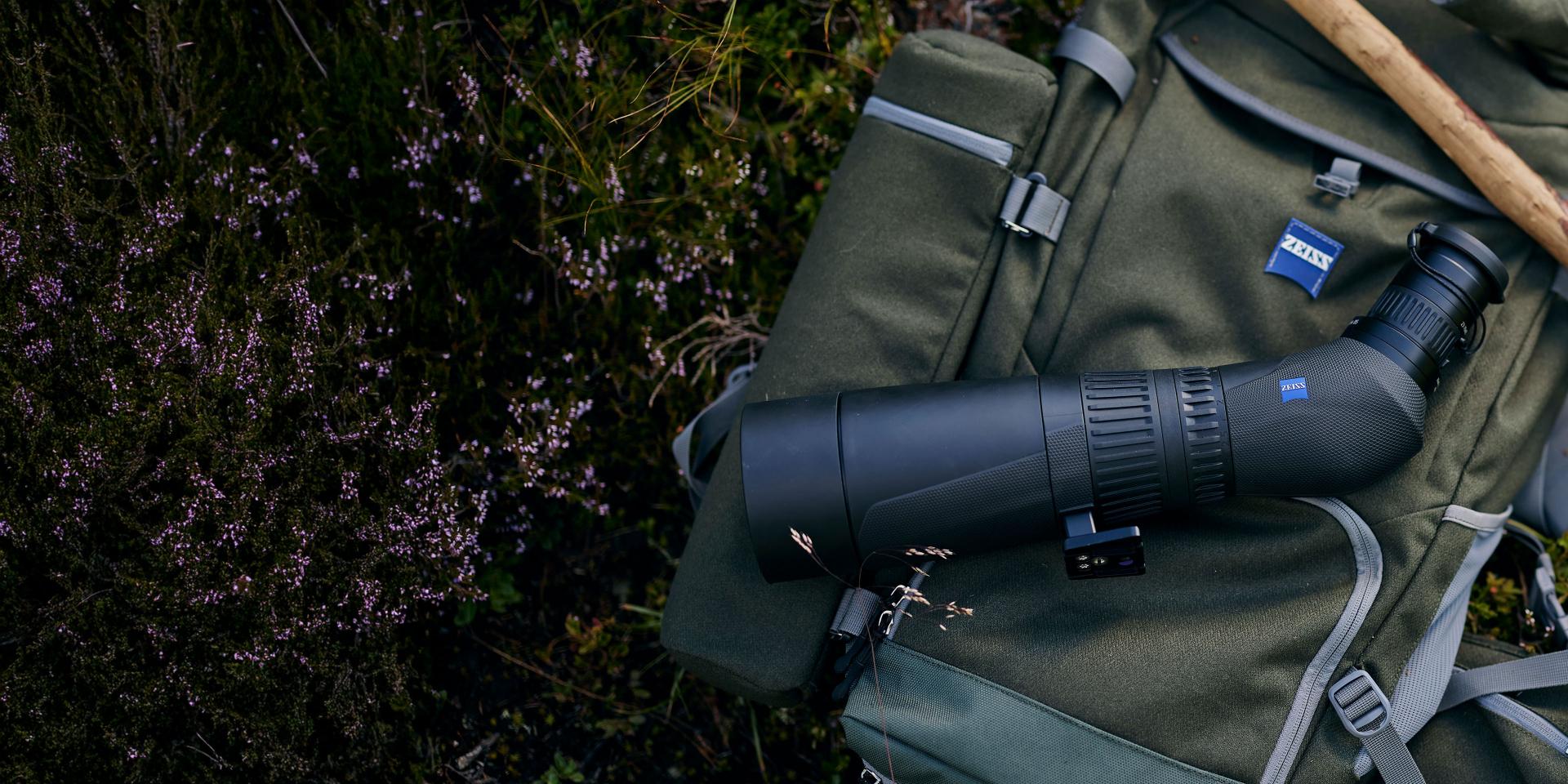 A ZEISS spotting scope rests on a green hiking backpack placed on the ground beside tall grass and heather.