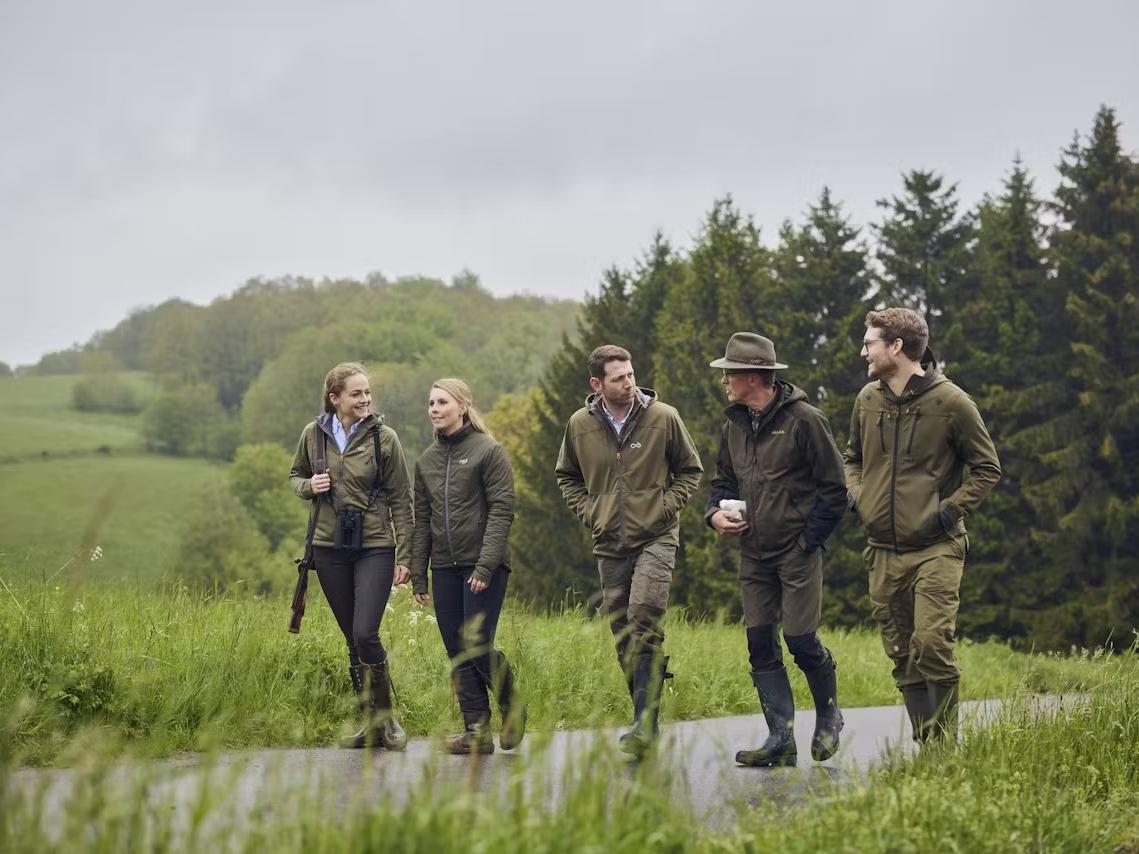 Eine Gruppe von jungen Jägerinnen und Jägern in Outdoor-Bekleidung spaziert gemeinsam durch eine grüne Landschaft, während sie Jagdausrüstung und ZEISS Ferngläser bei sich tragen.