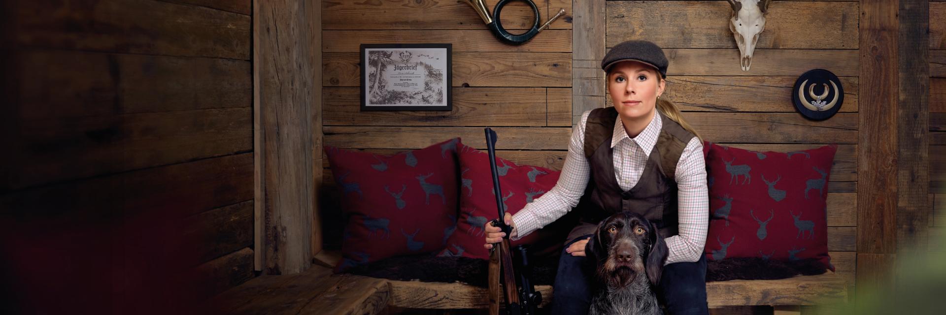 Eine Jägerin sitzt in einer rustikalen Holzhütte und hält ein Gewehr mit einem ZEISS Zielfernrohr in der Hand, begleitet von ihrem Jagdhund.