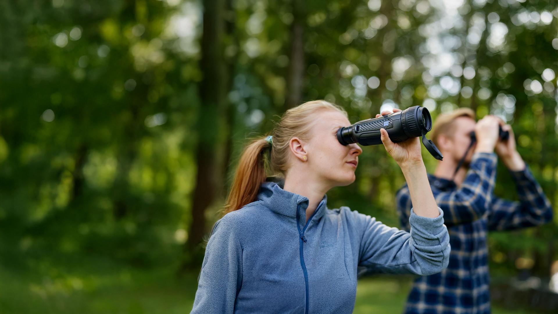 Two people are outdoors in a forested area using ZEISS optical devices for observation.