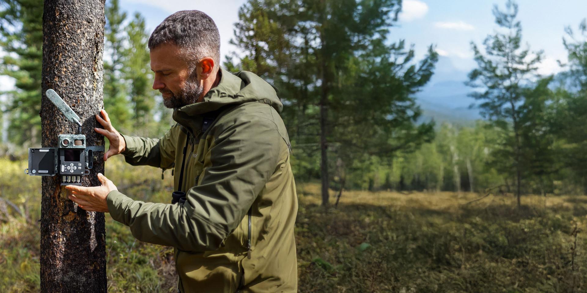 A man in outdoor gear mounts a ZEISS camera trap to a tree in a forested landscape.