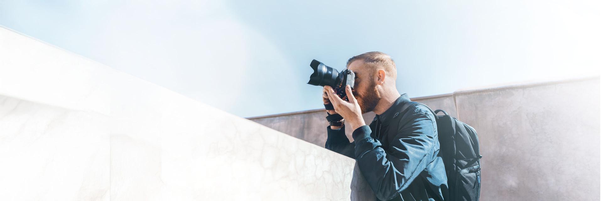 A man with a backpack is taking a photo with a camera equipped with a ZEISS lens in an outdoor urban setting.