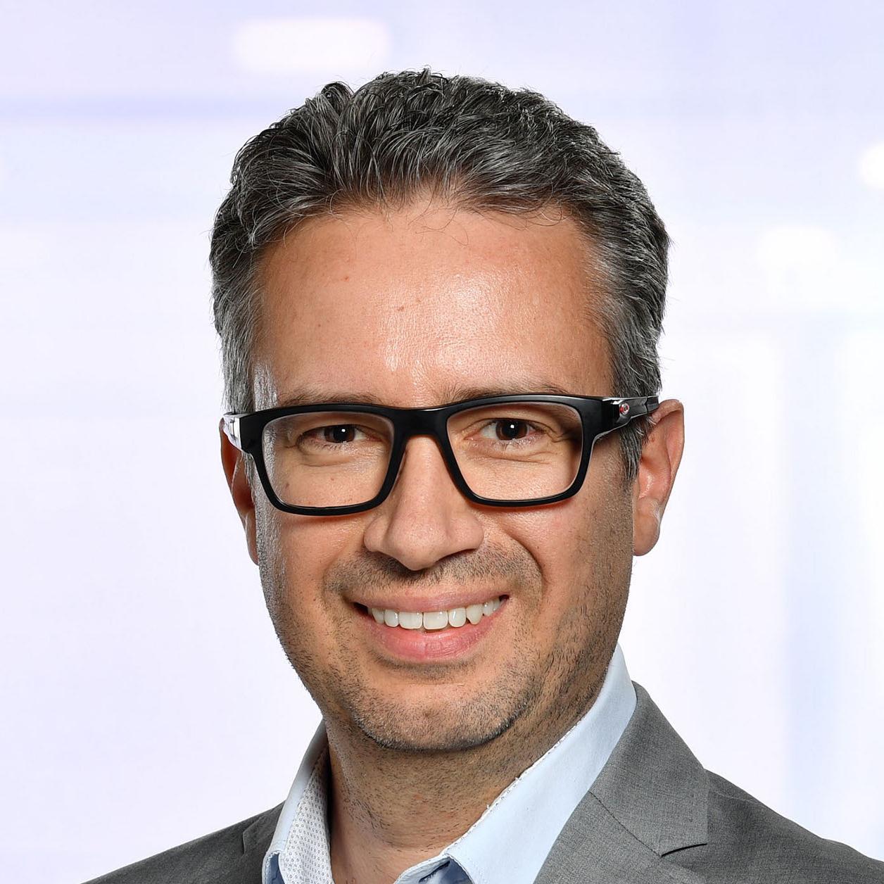 Portrait of a smiling man wearing glasses, a light blue shirt, and a grey blazer, standing in front of a softly blurred background, suitable for a professional ZEISS product website.