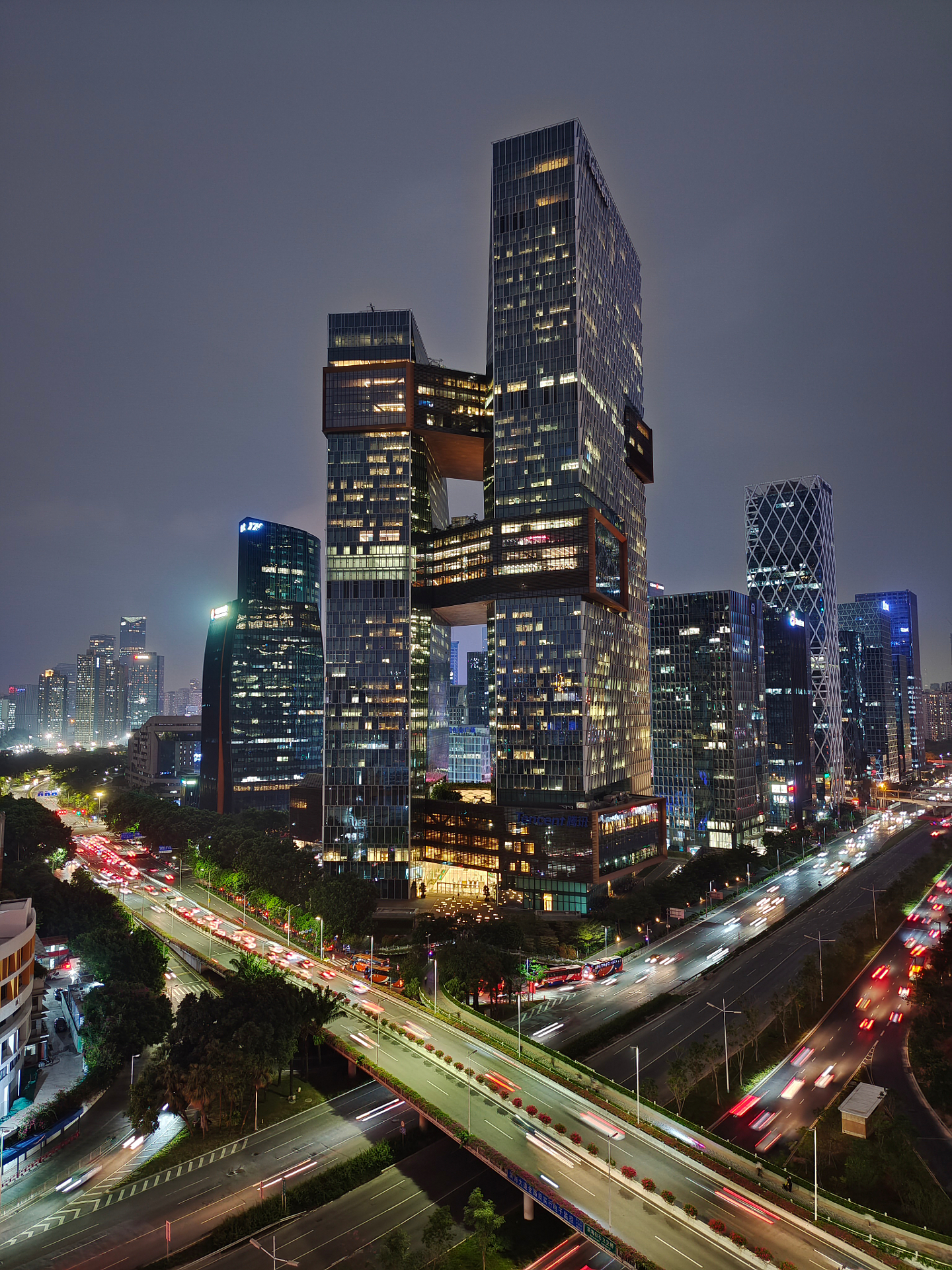 Modern cityscape at night featuring a tall, uniquely structured skyscraper illuminated with lights, surrounded by other high-rise buildings and busy traffic on multi-lane roads.