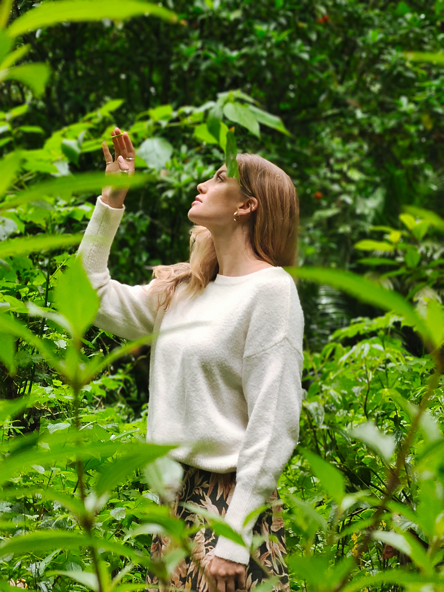 A woman wearing a white sweater stands in a lush green forest, looking upward while surrounded by dense foliage.