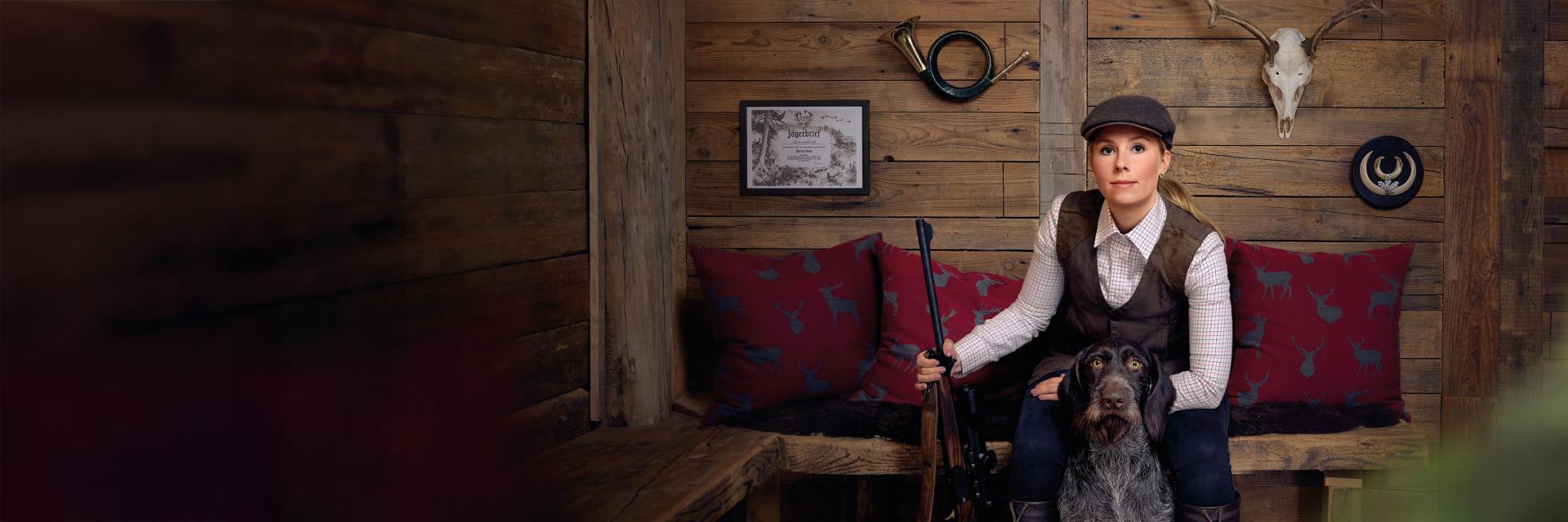 Eine Jägerin sitzt in einer rustikalen Holzhütte und hält ein Gewehr mit einem ZEISS Zielfernrohr in der Hand, begleitet von ihrem Jagdhund.