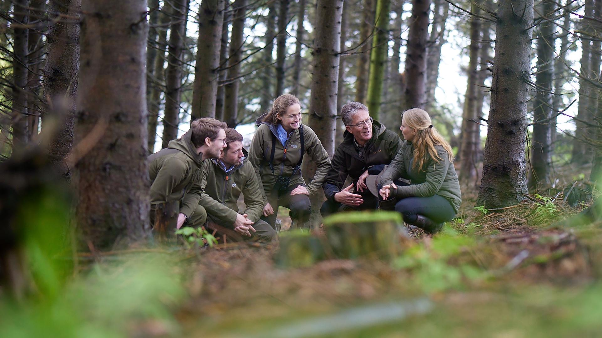 Eine Gruppe von Menschen in Outdoor-Kleidung nimmt an einem Waldseminar teil, unterhält sich und nutzt optische Geräte von ZEISS.