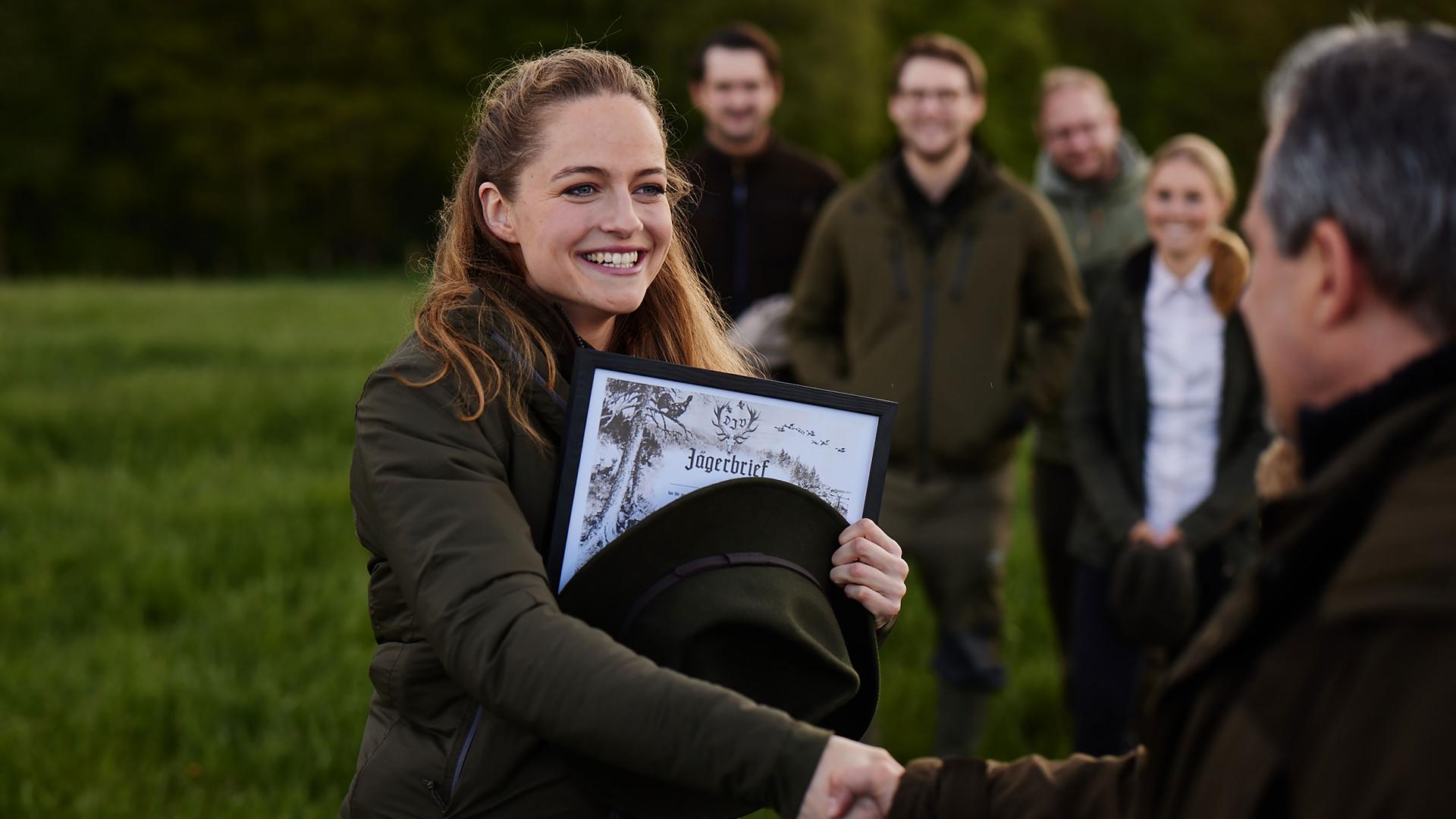 Eine lächelnde Frau erhält bei einer Zeremonie im Freien, umgeben von anderen Teilnehmern in ZEISS-Jagdbekleidung, mit einem Handschlag ihr Jägerbrief-Zertifikat.