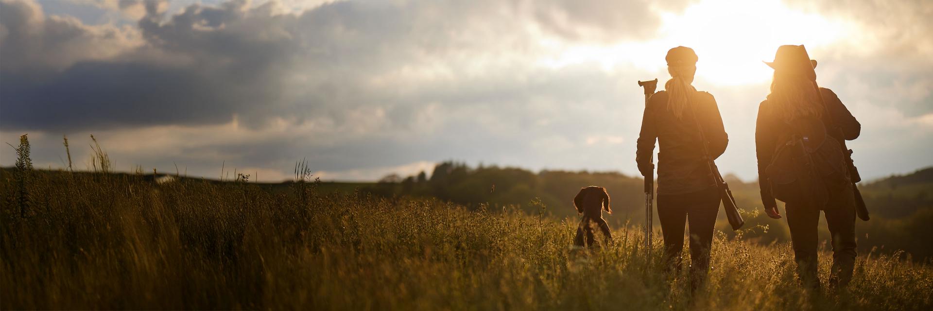 Zwei Jäger mit Gewehren und einem Jagdhund laufen mit ZEISS-Ausrüstung durch ein sonnenbeschienenes Feld in Richtung Horizont.
