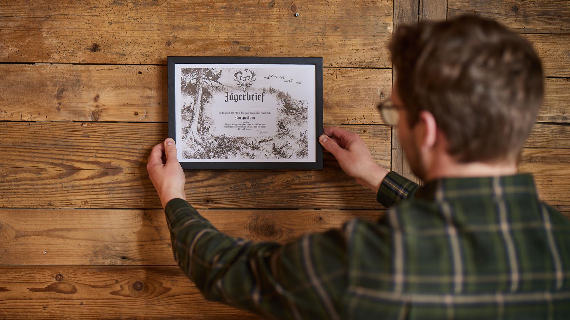 Eine Person in kariertem Hemd hält einen gerahmten Jägerbrief mit historischer Illustration und Schriftzug — detailreich und präzise abgebildet mit ZEISS Optik.