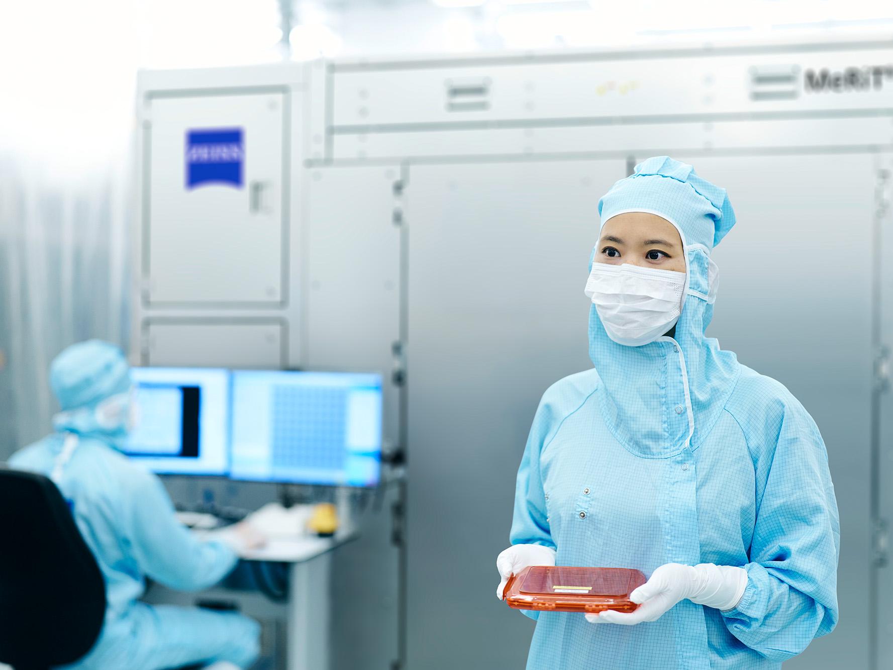 Eine Frau hält eine Maske im Reinraum der ZEISS SMT und im Hintergrund arbeitet ein Kollege am Bildschirm 