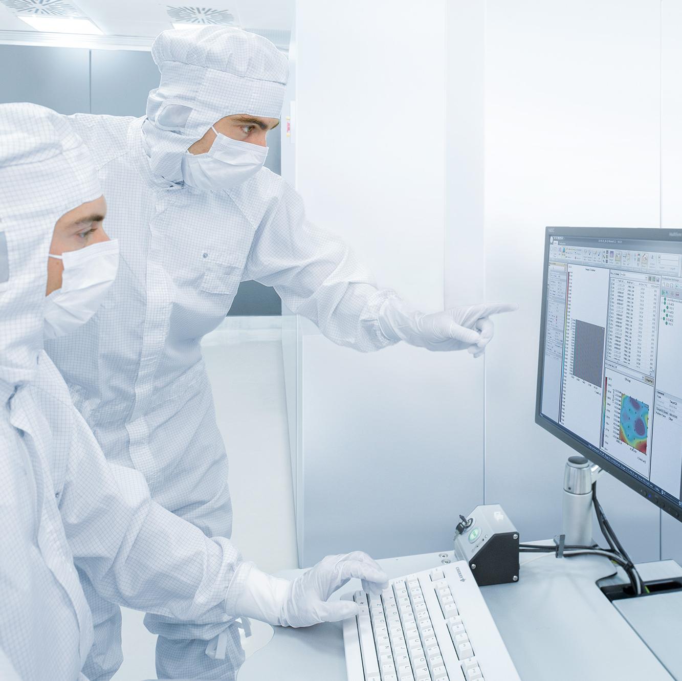 Zwei Mitarbeitende der ZEISS SMS arbeiten in Reimraumanzügen an einem Bildschirm-Arbeitsplatz.