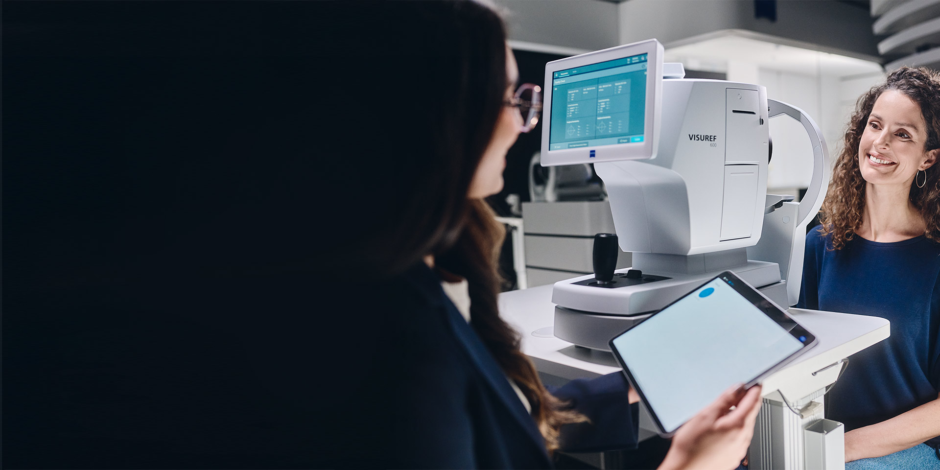 Eine Optikerin blickt auf ihr Tablet. Sie sitzt vor einem ZEISS VISUREF 600 und spricht mit einem Kunden.