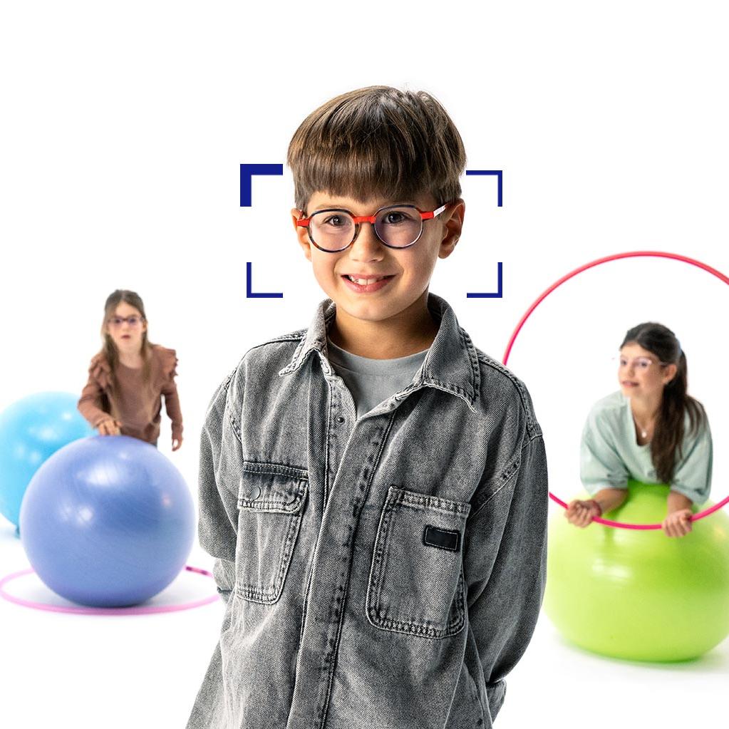 Ein Junge mit braunen Haaren, runder Brille und ZEISS MyoCare Brillengläsern steht im Vordergrund und lächelt in die Kamera. Im Hintergrund sind zwei Mädchen mit ZEISS MyoCare Brillengläsern zu sehen, die mit Reifen und Gymnastikbällen spielen.