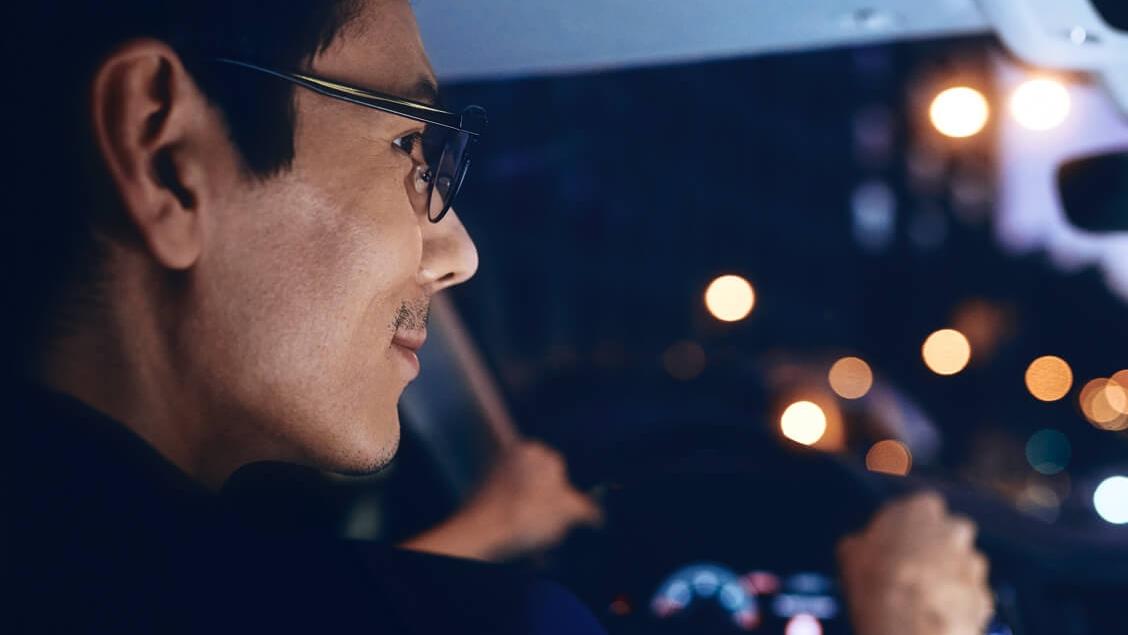 Mann mit Brille im Auto fährt durch die Dunkelheit. Man sieht verschiedene verschwommene Lichtquellen.