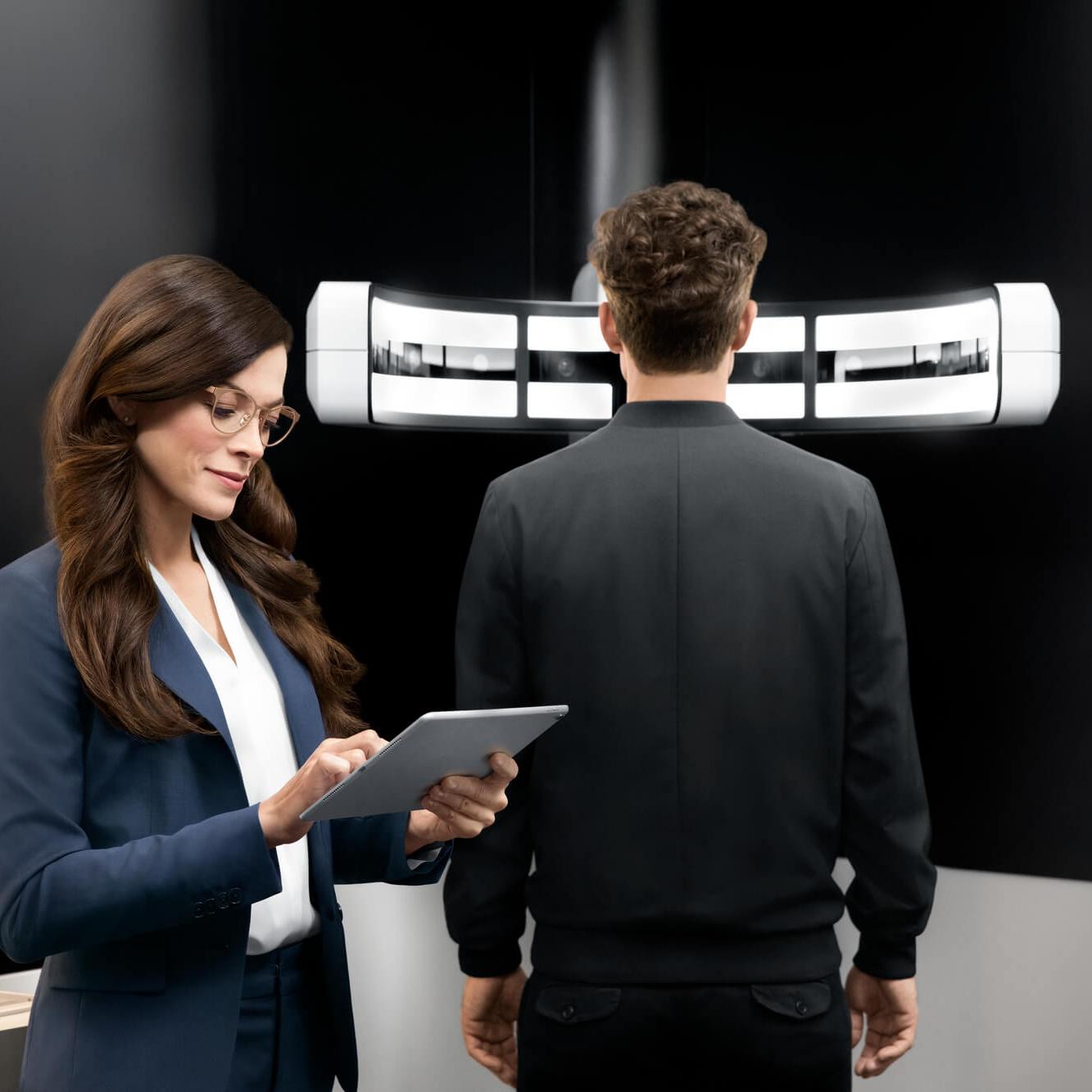 Eine Optikerin, die auf ein Tablet schaut und ein Mann der vor dem ZEISS VISUFIT 1000 steht.