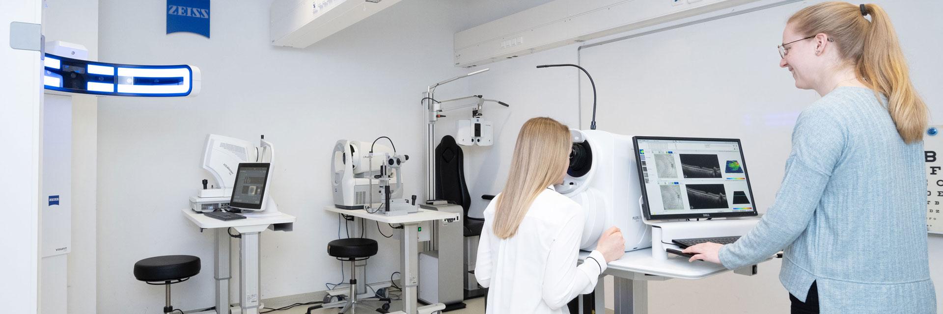Mitarbeiterin des Instituts schaut sich auf dem Display eines Messinstruments, an dem eine Probandin sitzt und durchschaut,  die Augenmessungen an.