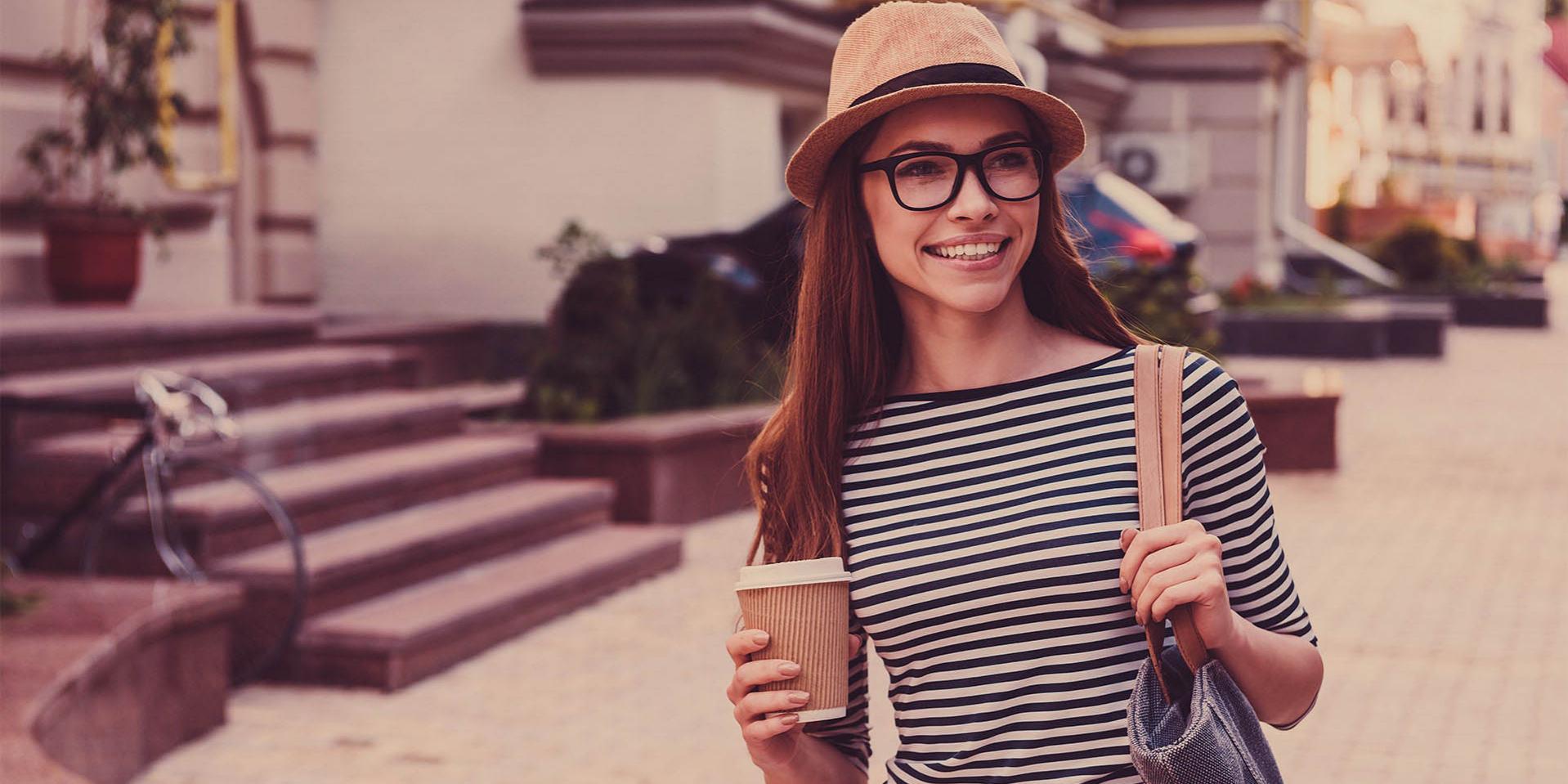 Eine junge Frau mit Mütze und Brille trägt einen Kaffeebecher (rechts) und eine Tasche auf der linken Schulter