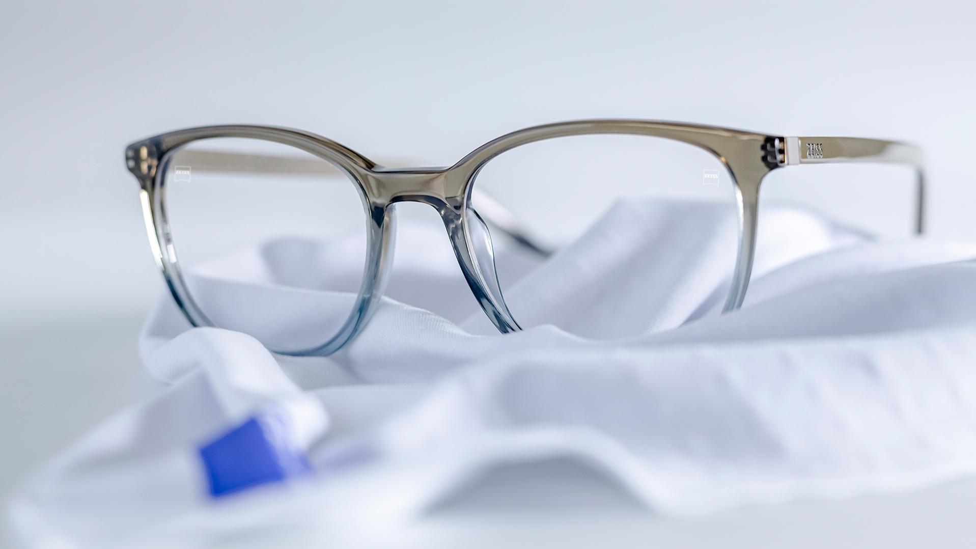 Eine Brille mit grau-blauer Fassung und ZEISS Brillengläsern mit DuraVision® Beschichtung liegt auf einem weißen Mikrofasertuch.