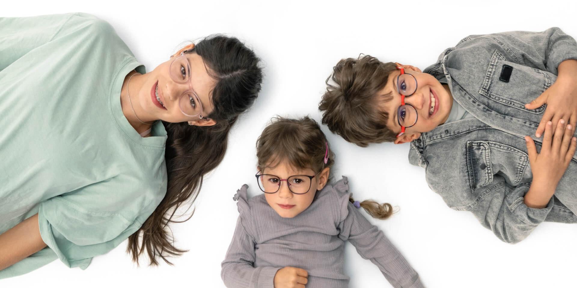 Eine Jugendliche und zwei jüngere Kinder mit ZEISS Brillengläsern zur Behandlung der Myopie liegen auf dem Boden.