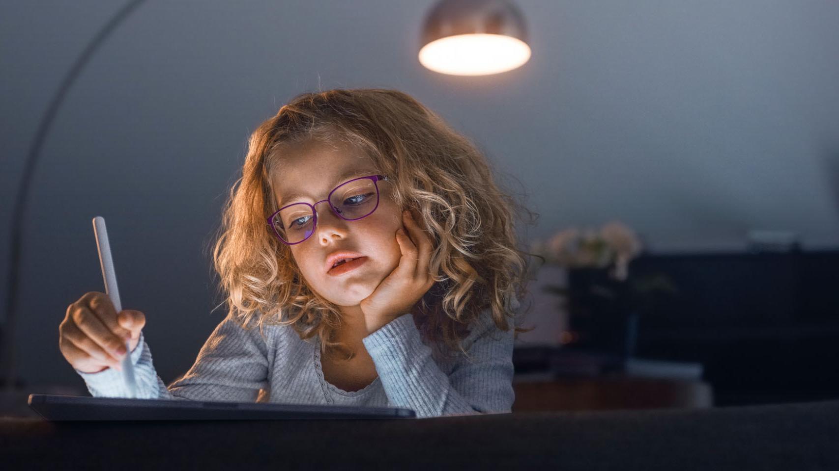 Ein kleines Mädchen in einem dunklen Wohnzimmer schaut auf ein Tablet.