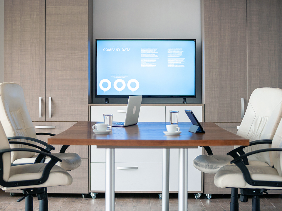 Sicht vom Büroschreibtisch einer Führungskraft. In einigen Metern Entfernung befindet sich ein Bildschirm an einer Wand. Es gibt einen kleinen Konferenztisch mit Bürostühlen, auf dem Tisch stehen Laptops.
