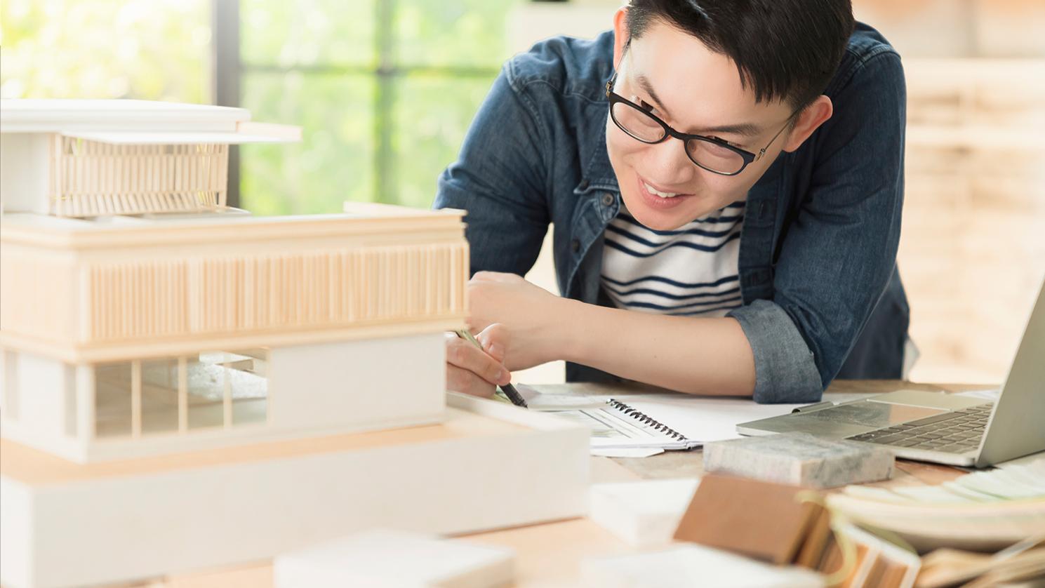 Ein Mann mittleren Alters mit kurzen schwarzen Haaren trägt eine ZEISS Brille für den Arbeitsplatz, die ihm die Fokussierung auf die Details seiner komplexen architektonischen Modelle erleichtert. 