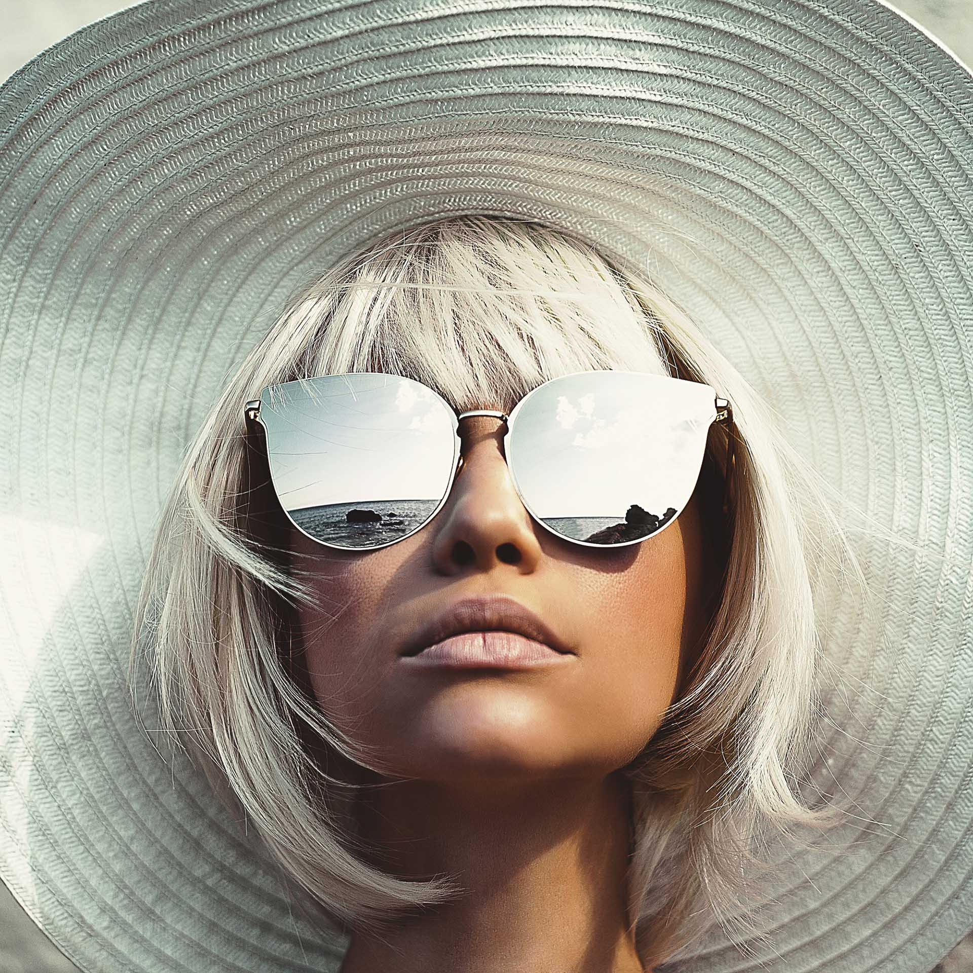 Eine junge Frau mit einem großen Sonnenhut und einer Sonnenbrille mit verspiegelten Brillengläsern schaut nach oben.