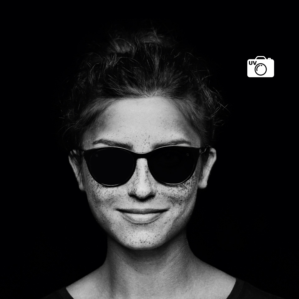 Ansicht derselben klare Brillengläser tragenden Frau durch eine normale Kamera und ein UV-Kameraobjektiv. Mit der UV-Kamera sieht das Brillenglas schwarz aus – ein Zeichen dafür, dass es keine UV-Strahlung durchlässt.  