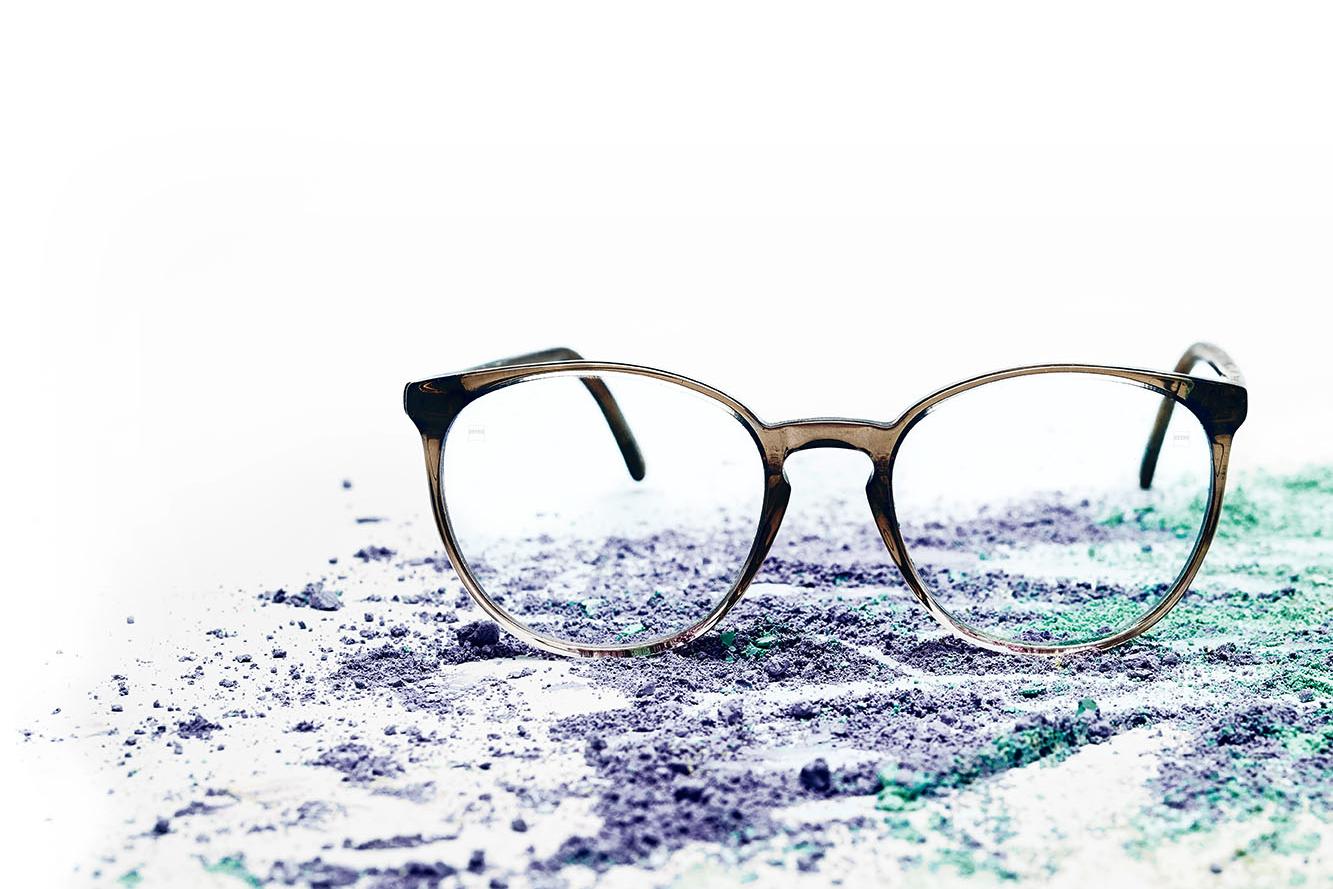Eine Brille mit klaren Brillengläsern liegt auf farbigem Puder.