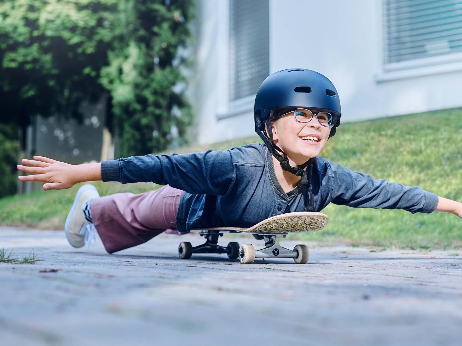 Ein Mädchen mit Helm und Brille rollt auf einem Skateboard liegend die Straße entlang und streckt die Arme aus.