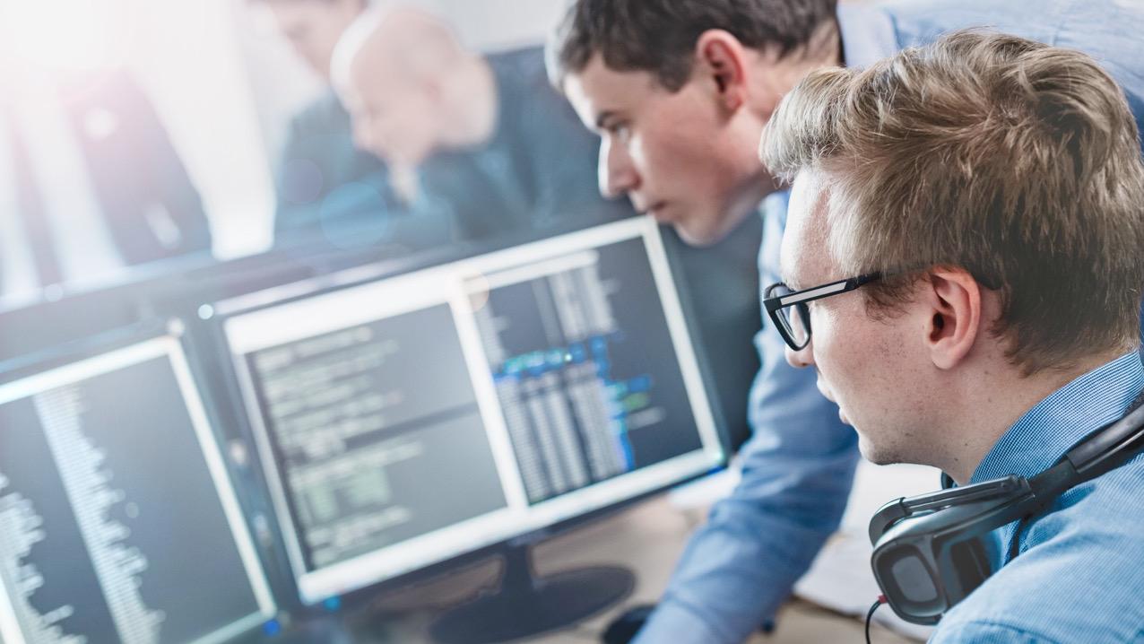 Zwei Softwareentwickler arbeiten zusammen, während sie in einer modernen Büroumgebung Code auf mehreren Computerbildschirmen analysieren.