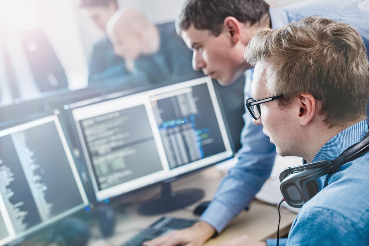 Zwei Softwareentwickler arbeiten zusammen, während sie in einer modernen Büroumgebung Code auf mehreren Computerbildschirmen analysieren.