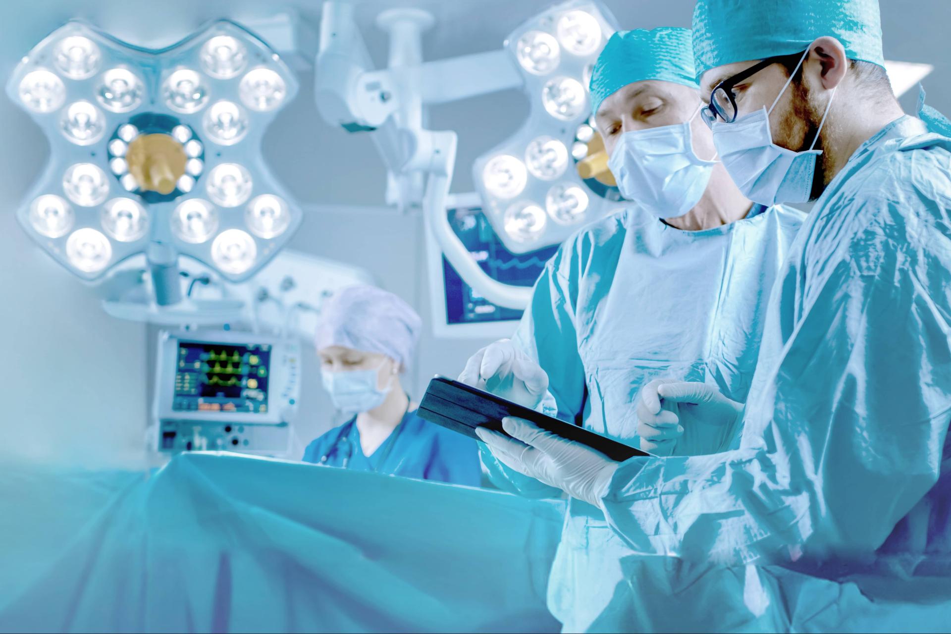 Chirurgen in einem hell erleuchteten Operationssaal tragen chirurgische Kittel und Masken, während sie Daten auf einem Tablet besprechen, wobei medizinische Geräte im Hintergrund zu sehen sind.